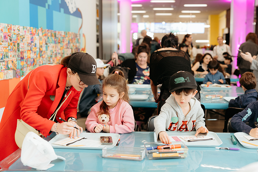 Adults and children sit at tables covered with art supplies, drawing and coloring. A woman in a red coat helps a young girl with brown hair, while a boy with a cap colors nearby. The room is busy and brightly lit.