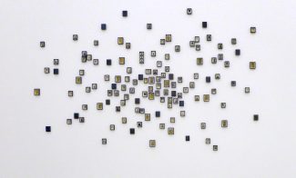 A white wall displays clusters of small, square tiles arranged in a loose, organic pattern, each tile containing simple black and white illustrations or symbols, with some yellow accents.