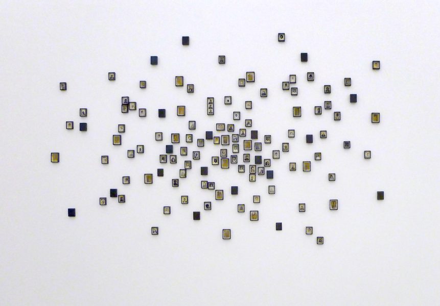 A white wall displays clusters of small, square tiles arranged in a loose, organic pattern, each tile containing simple black and white illustrations or symbols, with some yellow accents.