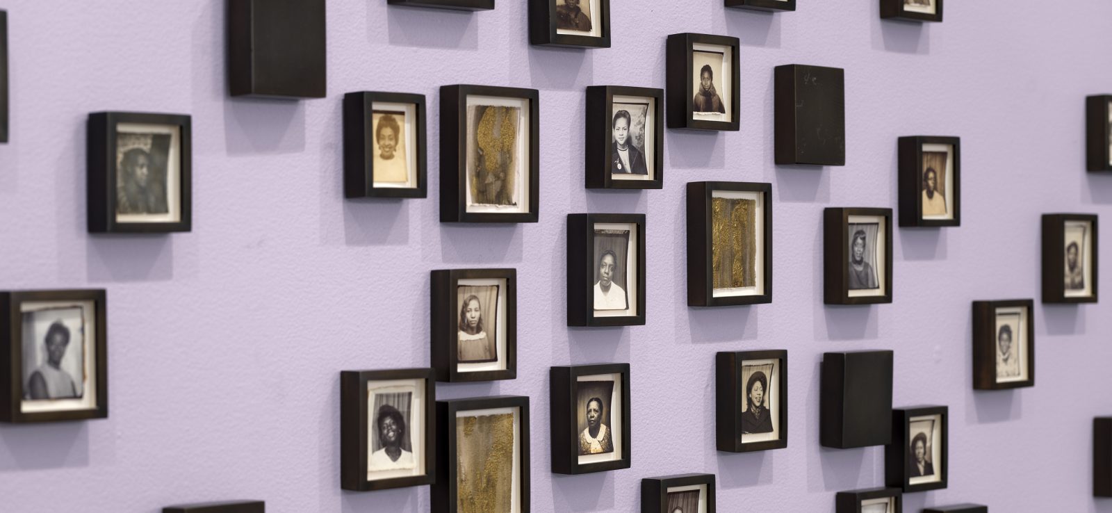 A lavender wall displays an arrangement of small, black-framed portraits in black and white—each featuring individual people