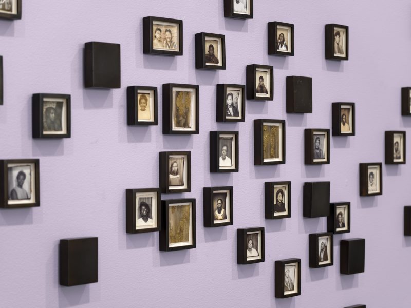 A lavender wall displays an arrangement of small, black-framed portraits in black and white—each featuring individual people