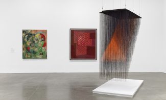 A gallery with two abstract paintings on a white wall and a large installation titled made of black and orange strings, suspended above a white platform. The space features a polished concrete floor.