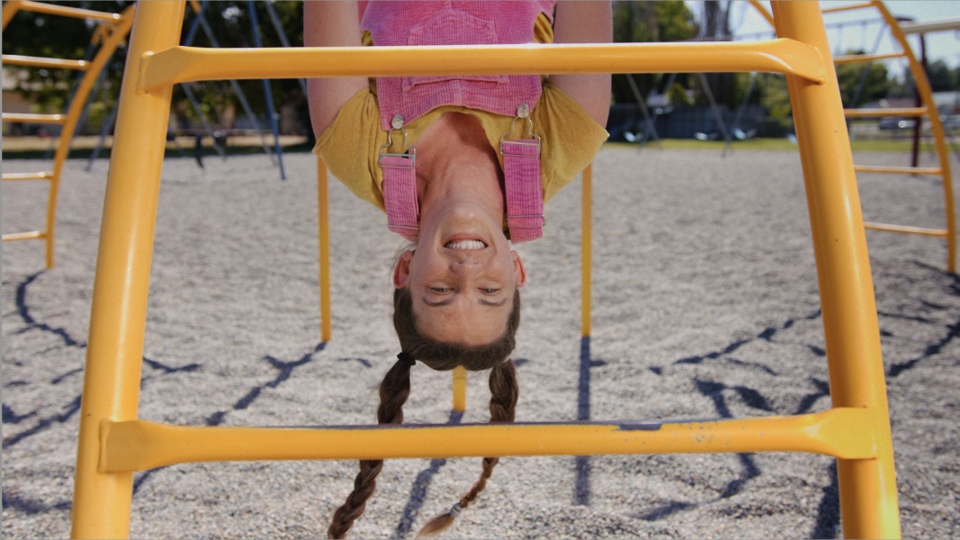 A young girl with braided hair hangs upside down from yellow playground bars, smiling and wearing pink overalls and a yellow shirt, with a gravel play area and park in the background.