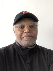A man wearing clear glasses, a black cap with a red P on it, and a black zip-up jacket stands in front of a plain white background, looking at the camera with a neutral expression.