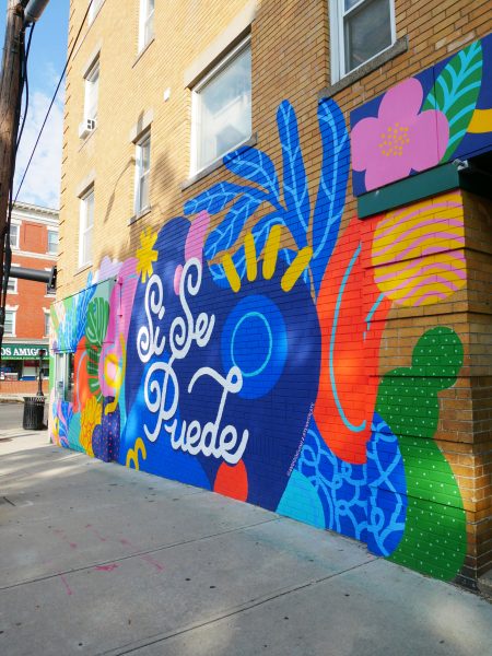 A colorful mural on a brick building features the phrase “Sí Se Puede” in white script, surrounded by vibrant floral and abstract patterns in blue, pink, yellow, orange, and green.