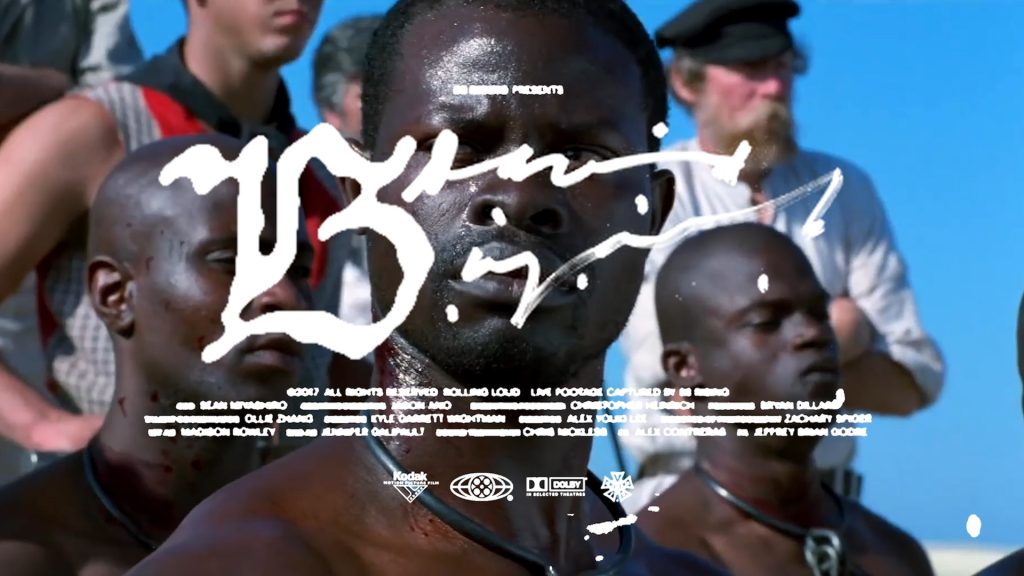 A close-up of a man with sweat on his face, standing among a group of people under bright sunlight. White handwritten text and film credits are overlaid across the image.