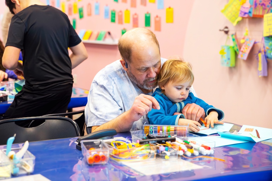 An older man helps a young child with a craft project at a table covered in art supplies, in a colorful room decorated with hanging tags and bright walls.