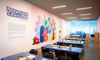 A bright, modern art classroom features tables set with art supplies, black chairs, and a vibrant mural of colorful flowers on the wall. Perpetual Gratitude and an artists statement are displayed beside the mural.