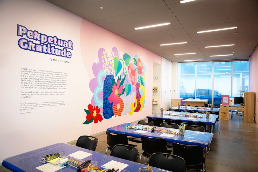 A bright, modern art classroom features tables set with art supplies, black chairs, and a vibrant mural of colorful flowers on the wall. Perpetual Gratitude and an artists statement are displayed beside the mural.