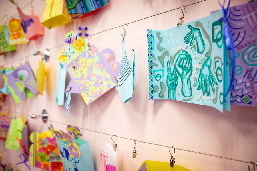 Colorful handmade cards and artwork hang on strings attached to a wall, each secured by clips. The cards feature bright drawings, patterns, and text, creating a lively and creative display.