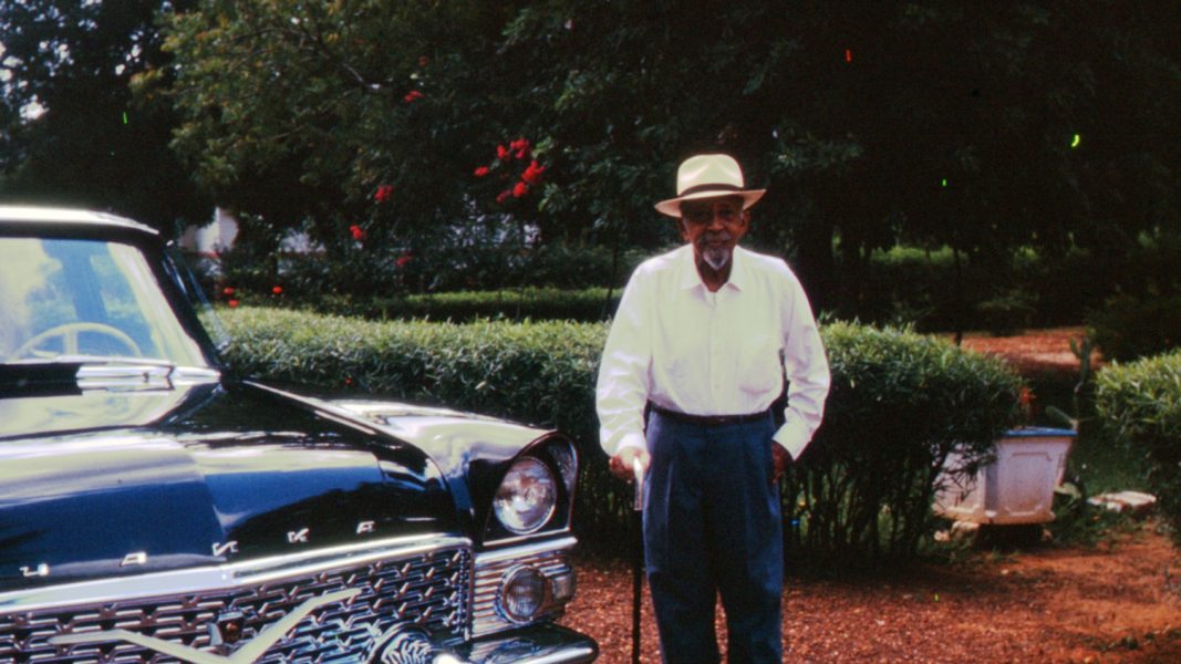 An elderly man wearing a hat, white shirt, and dark pants stands outdoors beside a vintage black car, holding a cane, with trees and bushes in the background.