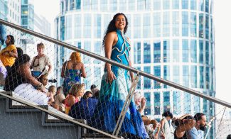 A woman in a vibrant blue and white dress stands confidently on outdoor stairs, surrounded by people sitting and chatting. Modern glass buildings and string lights are visible in the background.