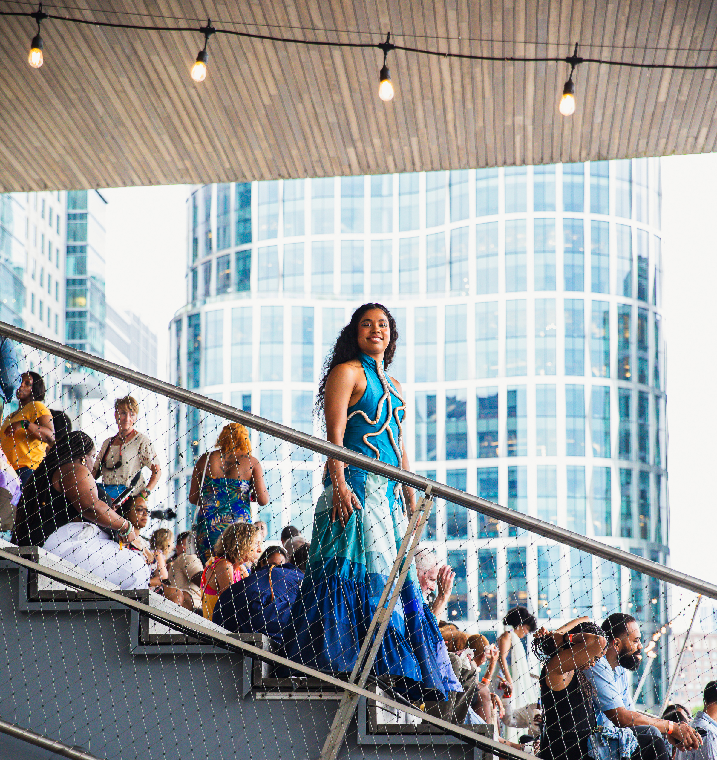 A woman in a vibrant blue and white dress stands confidently on outdoor stairs, surrounded by people sitting and chatting. Modern glass buildings and string lights are visible in the background.