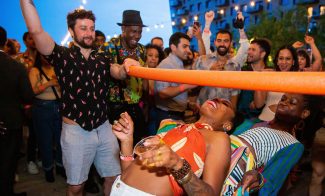 A lively group of people enjoy an outdoor party at dusk, cheering as two women limbo under an orange pool noodle. String lights glow above and everyone looks excited and festive.
