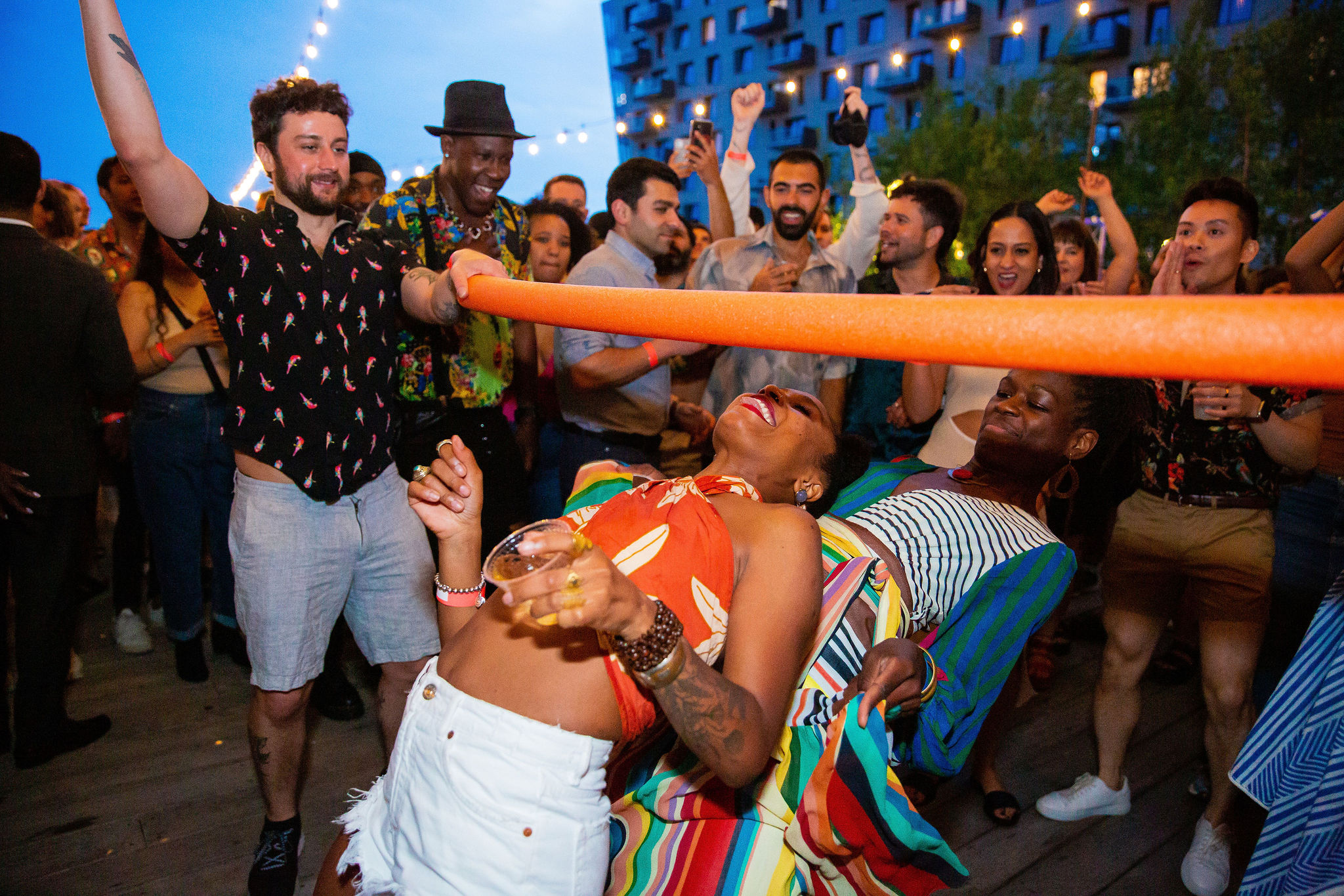A lively group of people enjoy an outdoor party at dusk, cheering as two women limbo under an orange pool noodle. String lights glow above and everyone looks excited and festive.