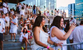 Two women in white dresses stand by a railing at an outdoor event. One smiles, while the other takes a photo with her phone. People in white attire and string lights decorate the steps in the background.