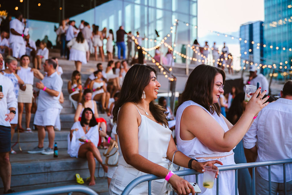 Two women in white dresses stand by a railing at an outdoor event. One smiles, while the other takes a photo with her phone. People in white attire and string lights decorate the steps in the background.