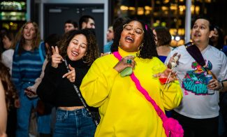 Two women dance and sing joyfully at a party; one wears a yellow outfit and holds drinks, the other is in a black top and jeans. People around them enjoy the lively atmosphere.