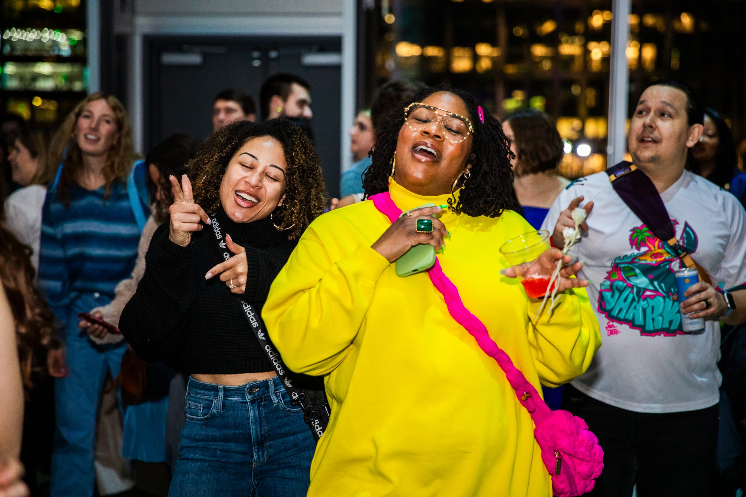 Two women dance and sing joyfully at a party; one wears a yellow outfit and holds drinks, the other is in a black top and jeans. People around them enjoy the lively atmosphere.