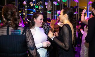 A group of people dressed in semi-formal attire mingle and chat at a lively indoor event with colorful lights, festive decorations, and a large window showing city buildings in the background.