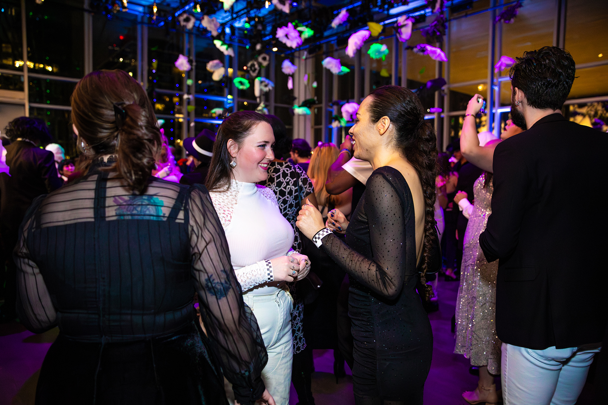 A group of people dressed in semi-formal attire mingle and chat at a lively indoor event with colorful lights, festive decorations, and a large window showing city buildings in the background.