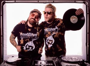 Two men in sunglasses and matching “Glitter Boys” shirts pose in front of DJ equipment. One holds a vinyl record while the other has his arm around his friend, both smiling against a bright white background.