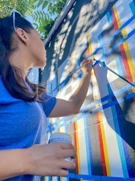 A woman in a blue shirt paints colorful lines on a wall mural outdoors, holding a brush in one hand and a jar in the other, with sunlight and tree shadows cast on the wall.