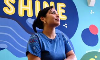 A woman in a blue shirt sits and looks up thoughtfully in front of a colorful mural that reads Together We Shine on a bright blue wall.