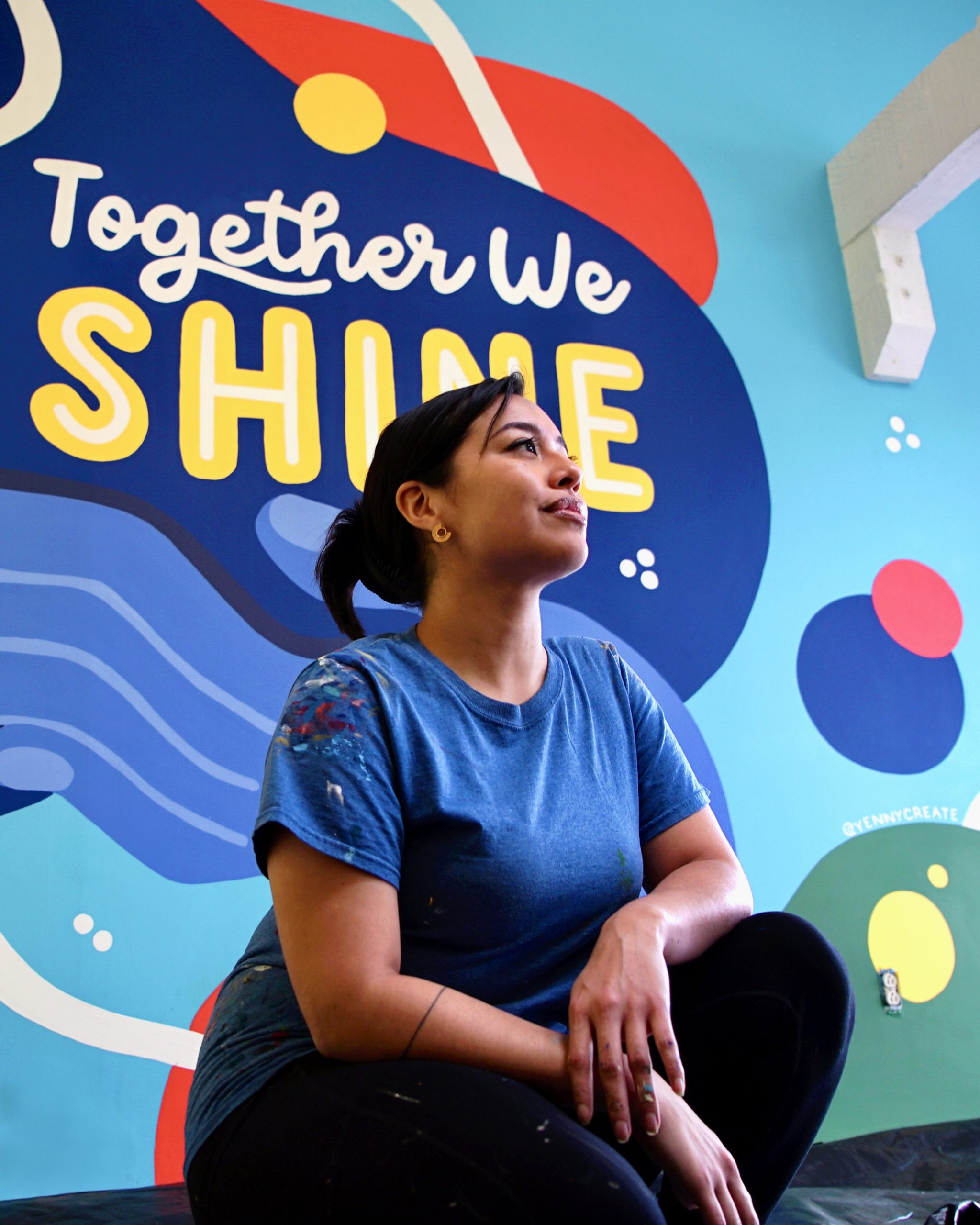 A woman in a blue shirt sits and looks up thoughtfully in front of a colorful mural that reads Together We Shine on a bright blue wall.