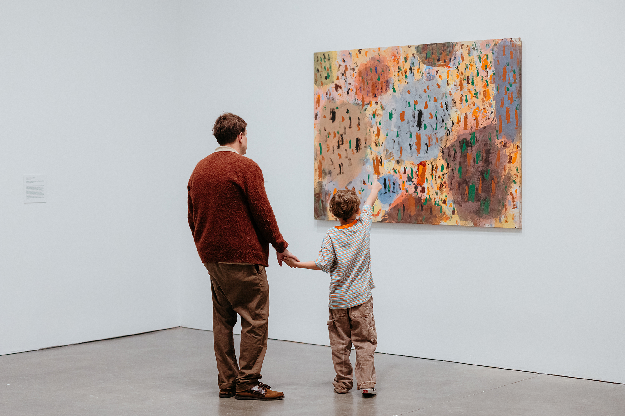 An adult and a child holding hands look at an abstract, colorful painting in an art gallery with white walls.