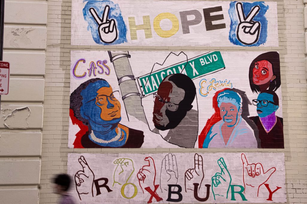 A mural on a brick wall features portraits of prominent Black figures, street signs reading Malcolm X Blvd, the word HOPE, peace signs, and ROXBURY spelled out in both letters and American Sign Language.