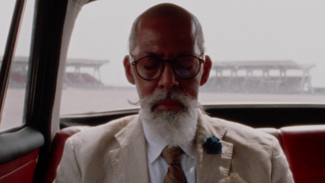 An older man with a white beard, mustache, glasses, and a bald head sits in a car with his eyes closed. He wears a light-colored suit, a patterned tie, and a blue lapel flower. The background shows blurred structures outside.