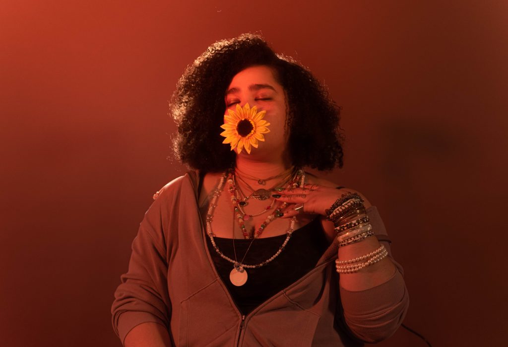 A person with curly hair and layered necklaces poses with a large yellow sunflower covering their mouth, eyes closed, against a warm orange-red background.