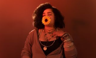 A person with curly hair and layered necklaces poses with a large yellow sunflower covering their mouth, eyes closed, against a warm orange-red background.