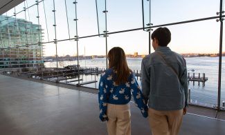 Two people holding hands walk inside a modern glass building, overlooking a waterfront with docks, boats, and a city skyline visible at sunset.