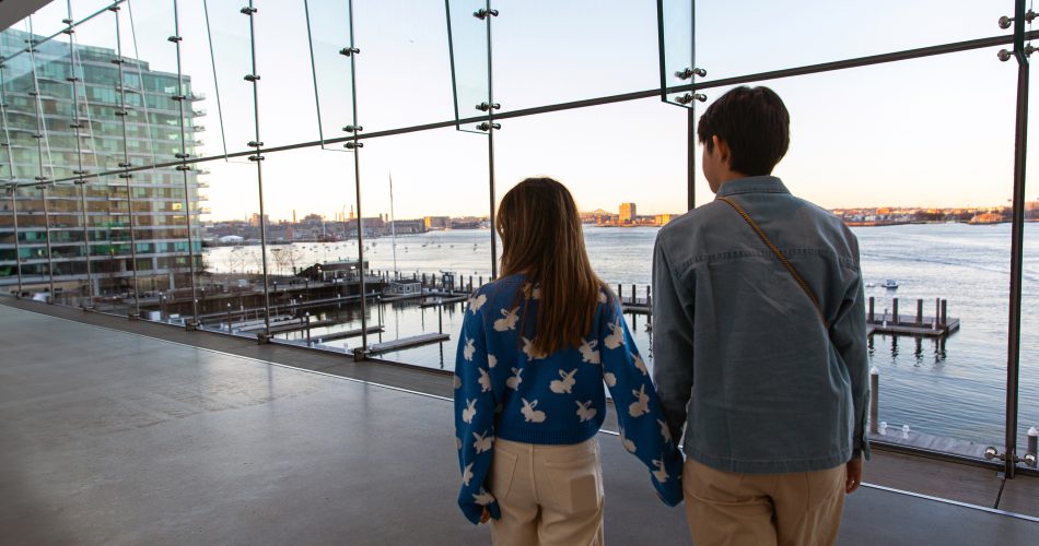 Two people holding hands walk inside a modern glass building, overlooking a waterfront with docks, boats, and a city skyline visible at sunset.