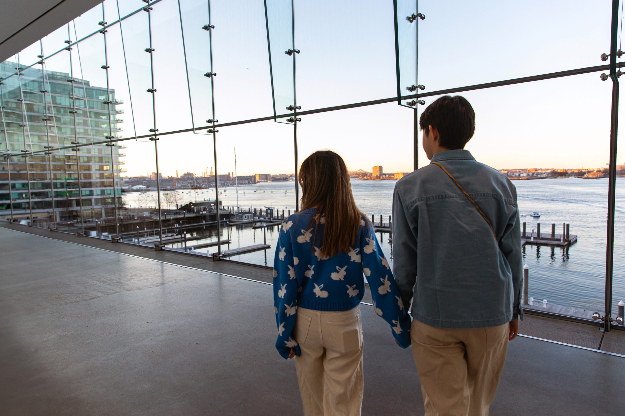 Two people holding hands walk inside a modern glass building, overlooking a waterfront with docks, boats, and a city skyline visible at sunset.