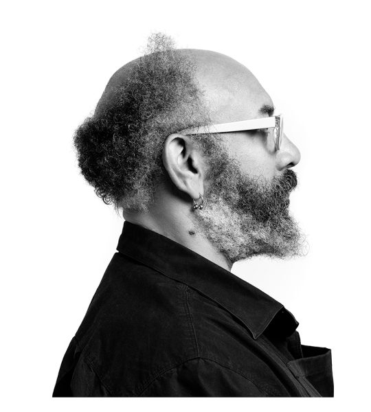 Black and white profile portrait of a bearded man with glasses, an earring, and a bald head with curly hair at the sides and back, wearing a collared dark shirt against a white background.