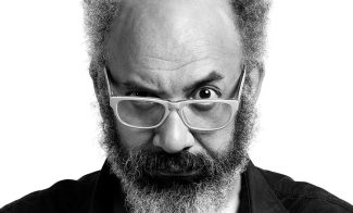 Black and white portrait of a man with a bald top, bushy beard, and unique hair styled into pointed tufts on each side. He wears glasses and has a slightly raised eyebrow, giving a curious expression.