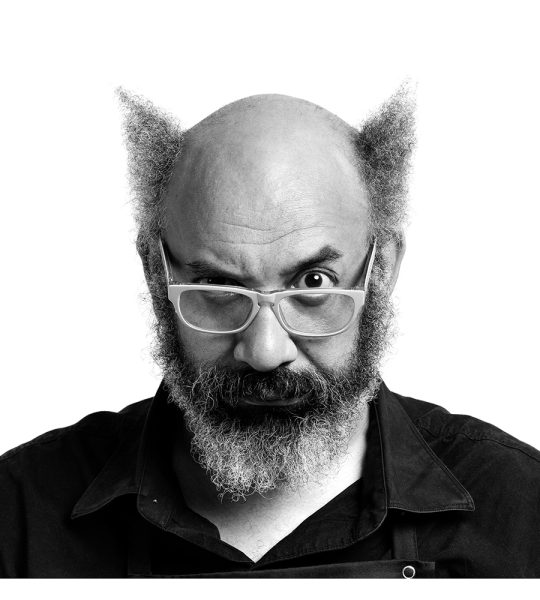 Black and white portrait of a man with a bald top, bushy beard, and unique hair styled into pointed tufts on each side. He wears glasses and has a slightly raised eyebrow, giving a curious expression.