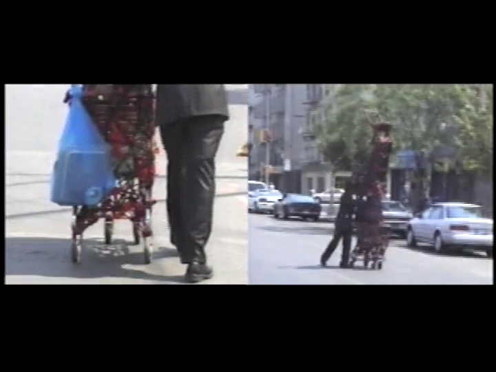 Two people walk on a city sidewalk, one pushing a cart with a large blue bag—evoking Nari Ward’s Pushing Savior. The split image offers two angles of the urban scene, surrounded by cars and buildings in the background.
