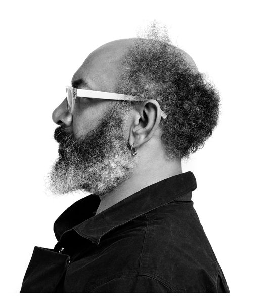 A black and white profile photo of a man with a beard and mustache, curly hair around the sides of his balding head, wearing glasses, a dark jacket, and a hoop earring, facing left against a white background.