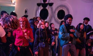 A diverse group of people, some smiling and dancing, stand together in a room with art with large circular patterns on the wall.