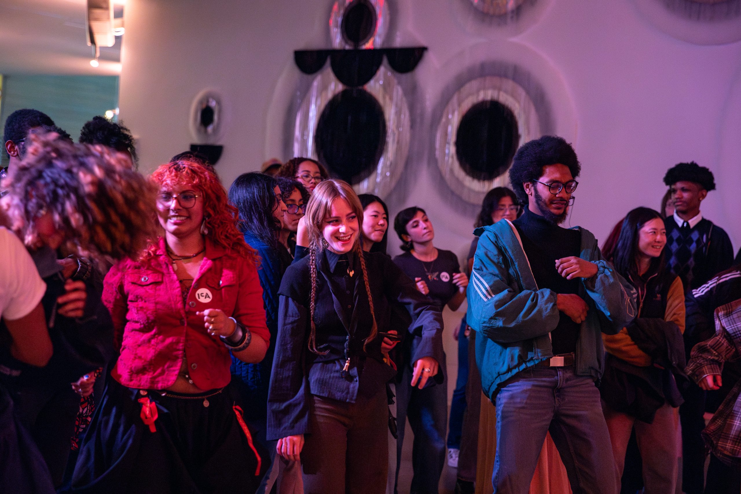 A diverse group of people, some smiling and dancing, stand together in a room with art with large circular patterns on the wall.