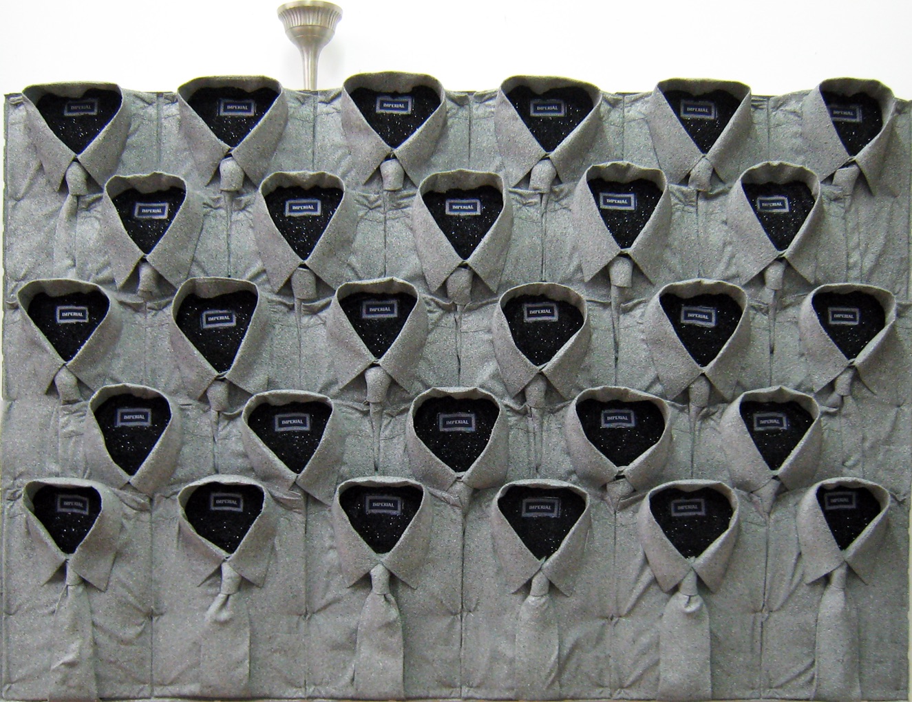 A wall display of 25 neatly arranged gray dress shirts, each buttoned up with the collars open and black fabric inside, giving the illusion of shirt fronts mounted on the wall. A light fixture is visible above.