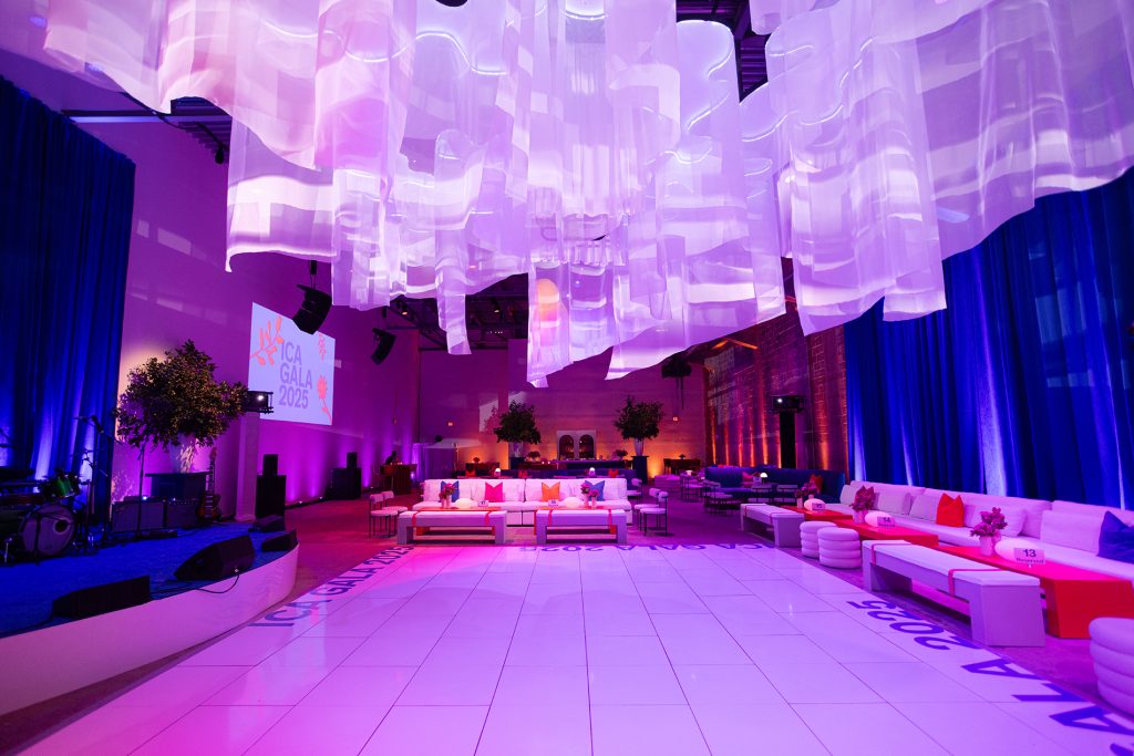 A modern event space with white couches, colorful pillows, blue uplighting, hanging translucent fabric decorations, and a stage with instruments. “ICA Gala 2025” is projected on the wall. The ambiance is vibrant and elegant.