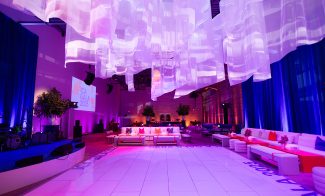 A modern event space with white couches, colorful pillows, blue uplighting, hanging translucent fabric decorations, and a stage with instruments. “ICA Gala 2025” is projected on the wall. The ambiance is vibrant and elegant.