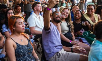 A group of people sitting closely together at an outdoor event. Several are smiling and posing for a selfie, while others hold drinks and chat, enjoying the lively atmosphere.