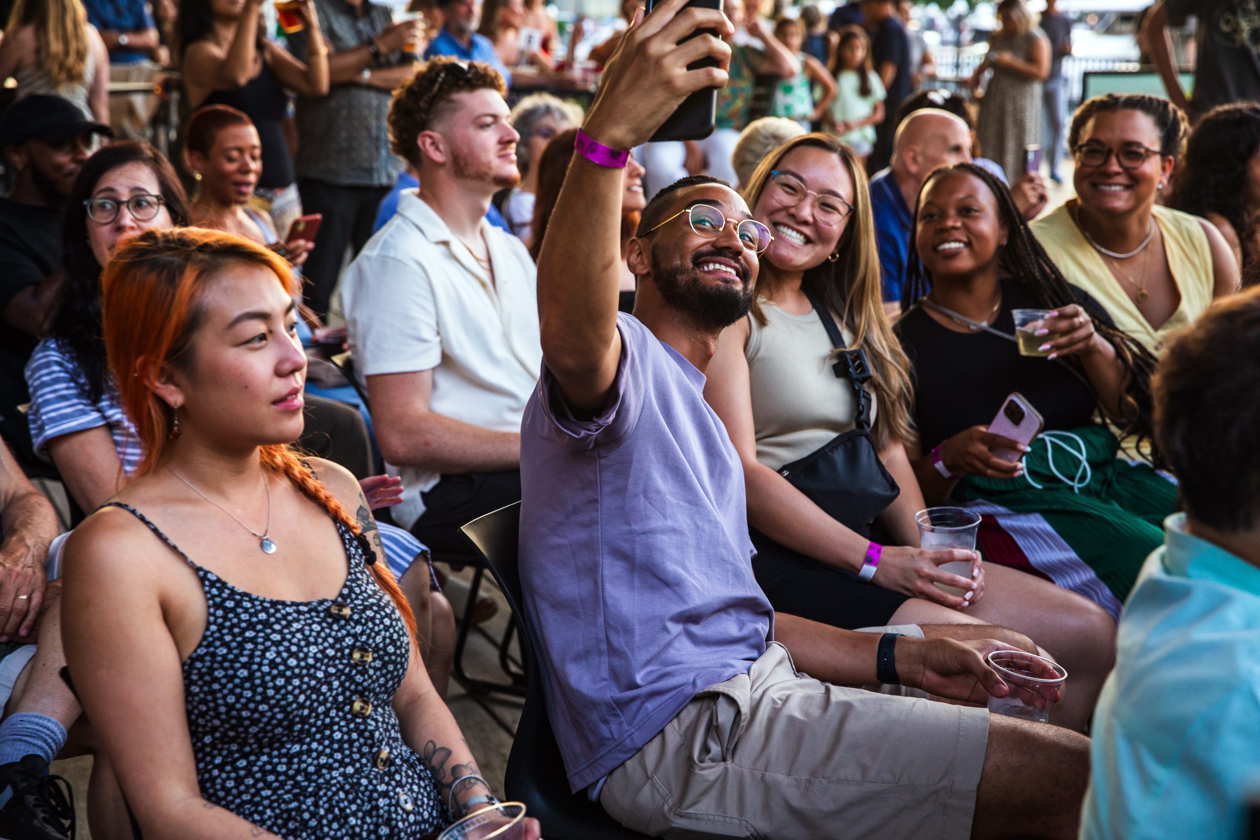 A group of people sitting closely together at an outdoor event. Several are smiling and posing for a selfie, while others hold drinks and chat, enjoying the lively atmosphere.
