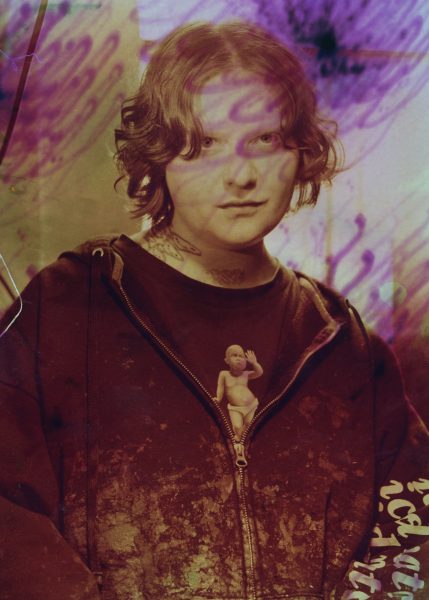 A person with wavy hair wearing a dark hoodie stands against a blurred, purple-toned background. A small doll figure appears attached to the hoodie, and abstract patterns overlay the image.